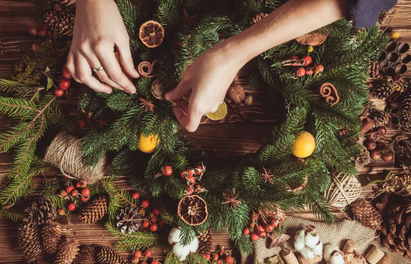 sapin-bio-fabrication_deco_artisanale Fabrication artisanale d’une couronne de Noël avec des branches de sapin, des fruits secs, des baies rouges et des éléments naturels sur une table en bois.